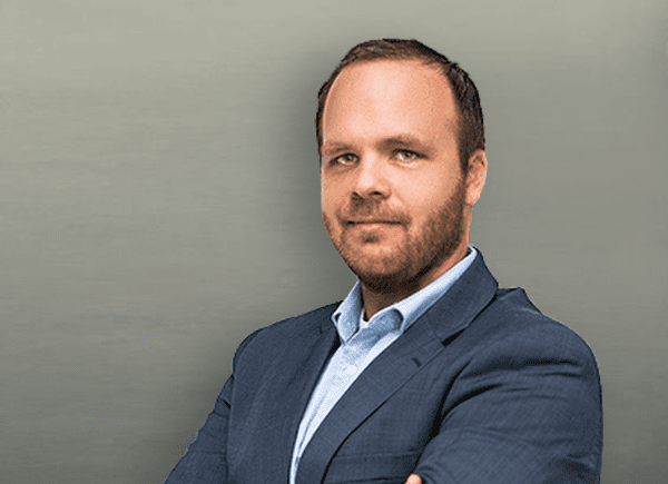 A man in a dark suit and light blue shirt stands with arms crossed, looking at the camera against a plain gray background.