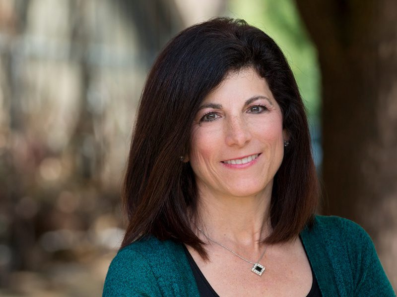 A woman with straight dark hair, wearing a green sweater and a necklace, smiles at the camera outdoors with a blurred background.