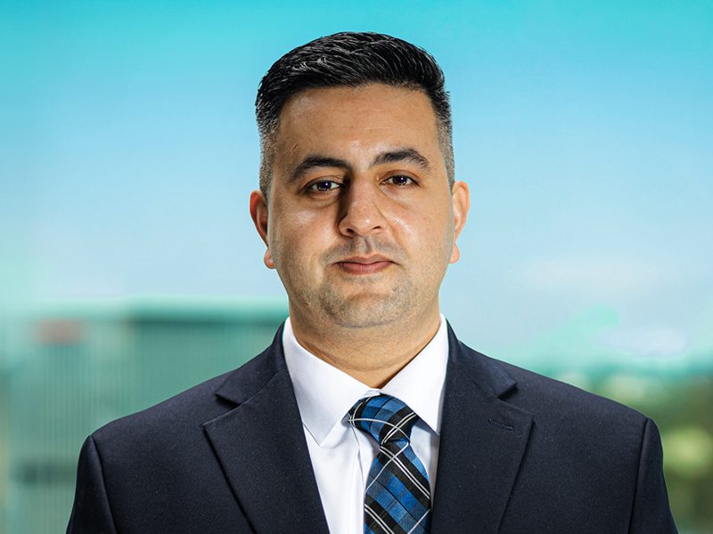 A man in a dark suit, white shirt, and blue plaid tie stands in front of a blurred outdoor background.