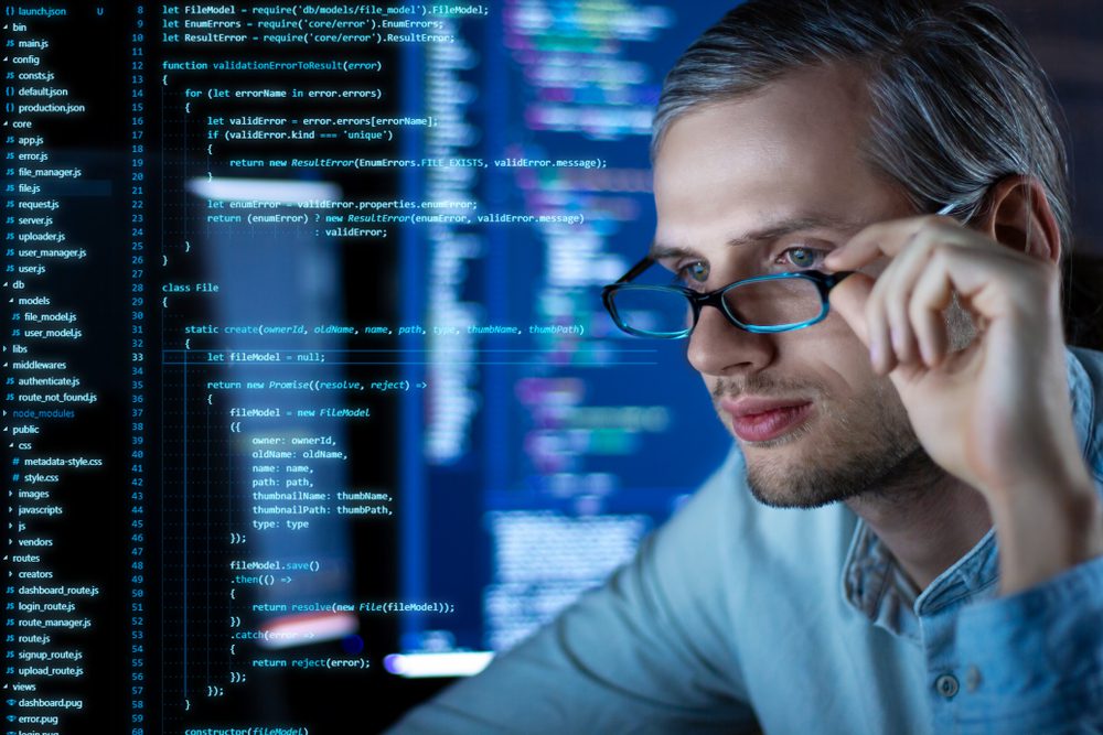 A person wearing glasses looks at computer screens displaying code and programming interfaces in a dimly lit workspace.