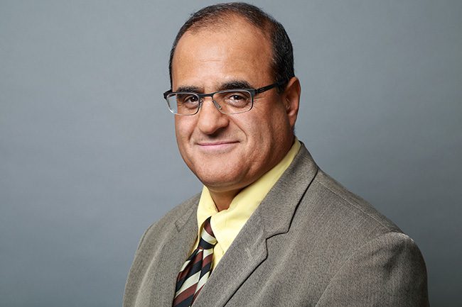 A middle-aged man wearing glasses, a gray suit, a yellow shirt, and a striped tie poses in front of a plain gray background.