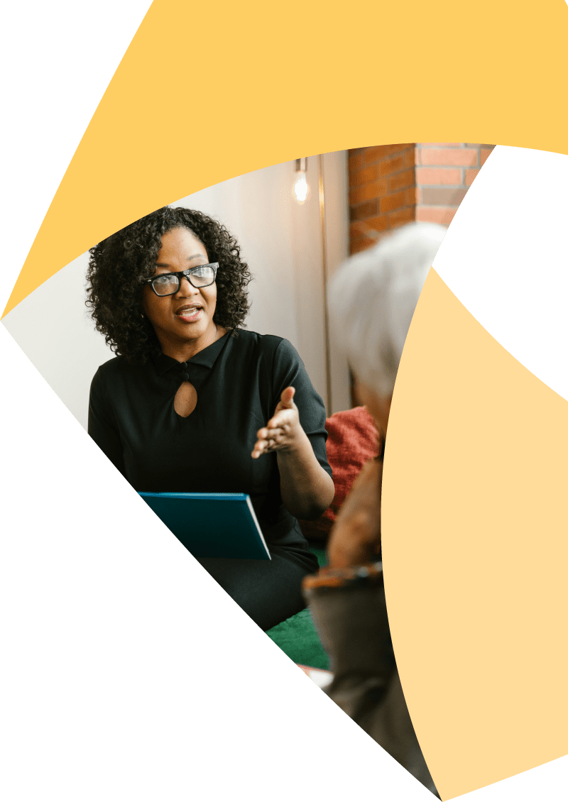 A woman with curly hair speaks while holding a folder, gesturing with her hand toward another seated person. Geometric yellow shapes frame the image.