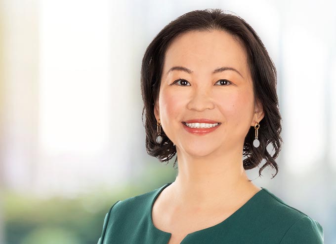 A woman with short dark hair, wearing a green top and drop earrings, smiles at the camera against a softly blurred background.