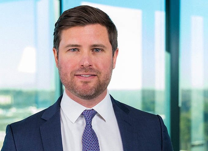 A man in a navy suit and patterned tie stands in front of large glass windows with a cityscape in the background.