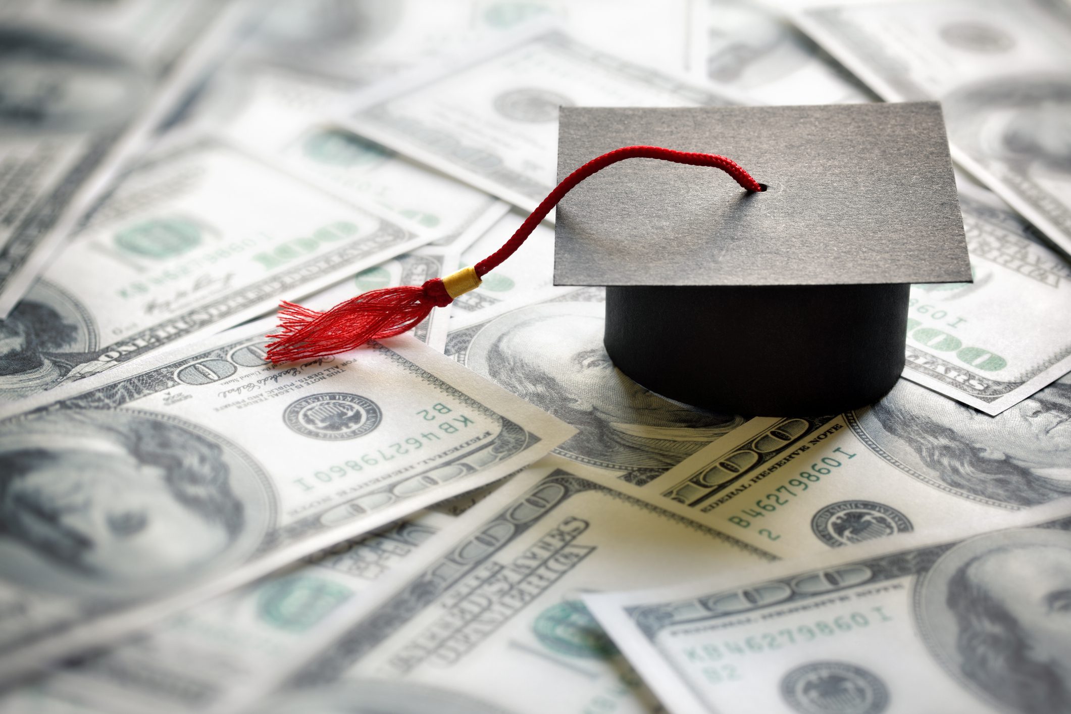 A graduation cap with a red tassel rests on a pile of one hundred dollar bills, symbolizing the cost of education or student loans.
