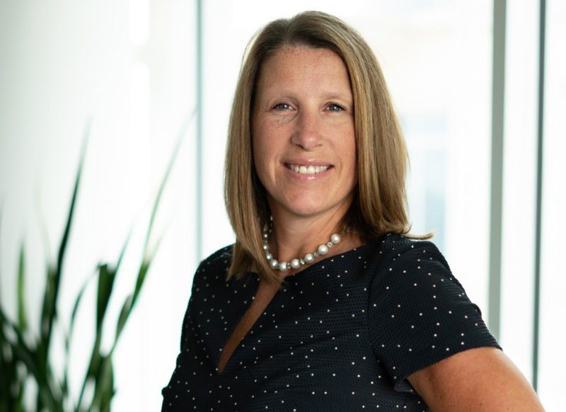 A woman with straight, shoulder-length hair wearing a black polka dot top and a pearl necklace smiles at the camera. A green plant is in the blurred background.