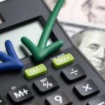 Calculator with TAX+ and TAX- buttons, a blue down arrow, a green up arrow, and part of a U.S. hundred-dollar bill in the background.