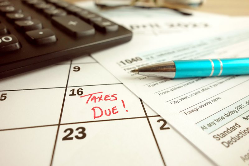 A calendar marked Taxes Due on the 16th, a tax form, a pen, and a calculator are shown on a desk.