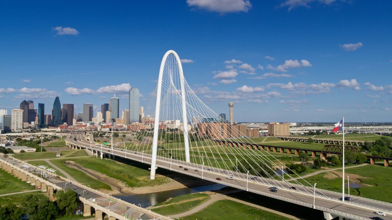 A modern cable-stayed bridge spans a river with city skyscrapers in the background and a large Texas flag on the right side.