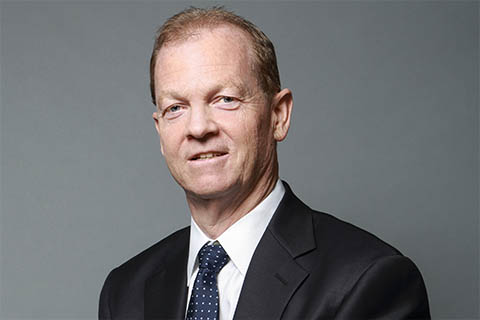 A man in a dark suit and polka dot tie poses in front of a plain gray background, facing the camera with a neutral expression.