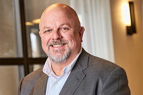 A middle-aged man with a bald head and goatee, wearing a gray blazer and white shirt, smiles at the camera indoors with soft lighting.