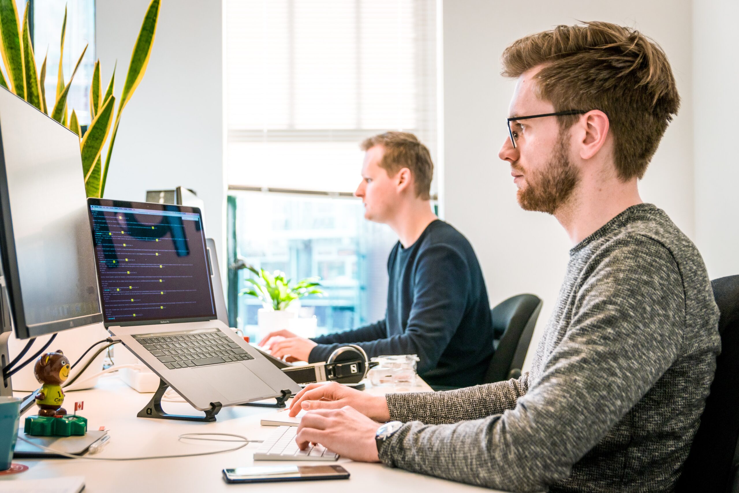 Two men work at desks with computers in a bright office. One uses a laptop on a stand and an external keyboard; the other is focused on his desktop monitor.