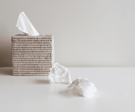 A woven tissue box sits on a white surface with two crumpled tissues lying beside it.