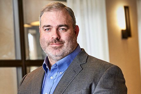 A man with short gray hair and a beard wearing a blue shirt and gray blazer stands indoors in front of windows and a wall sconce.