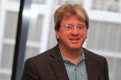 A middle-aged man with glasses and light brown hair wearing a checkered shirt and dark blazer, standing indoors in front of large windows.