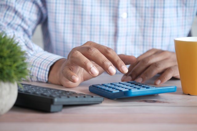 A person uses a blue calculator on a desk, with a keyboard, a potted plant, and a yellow cup nearby.