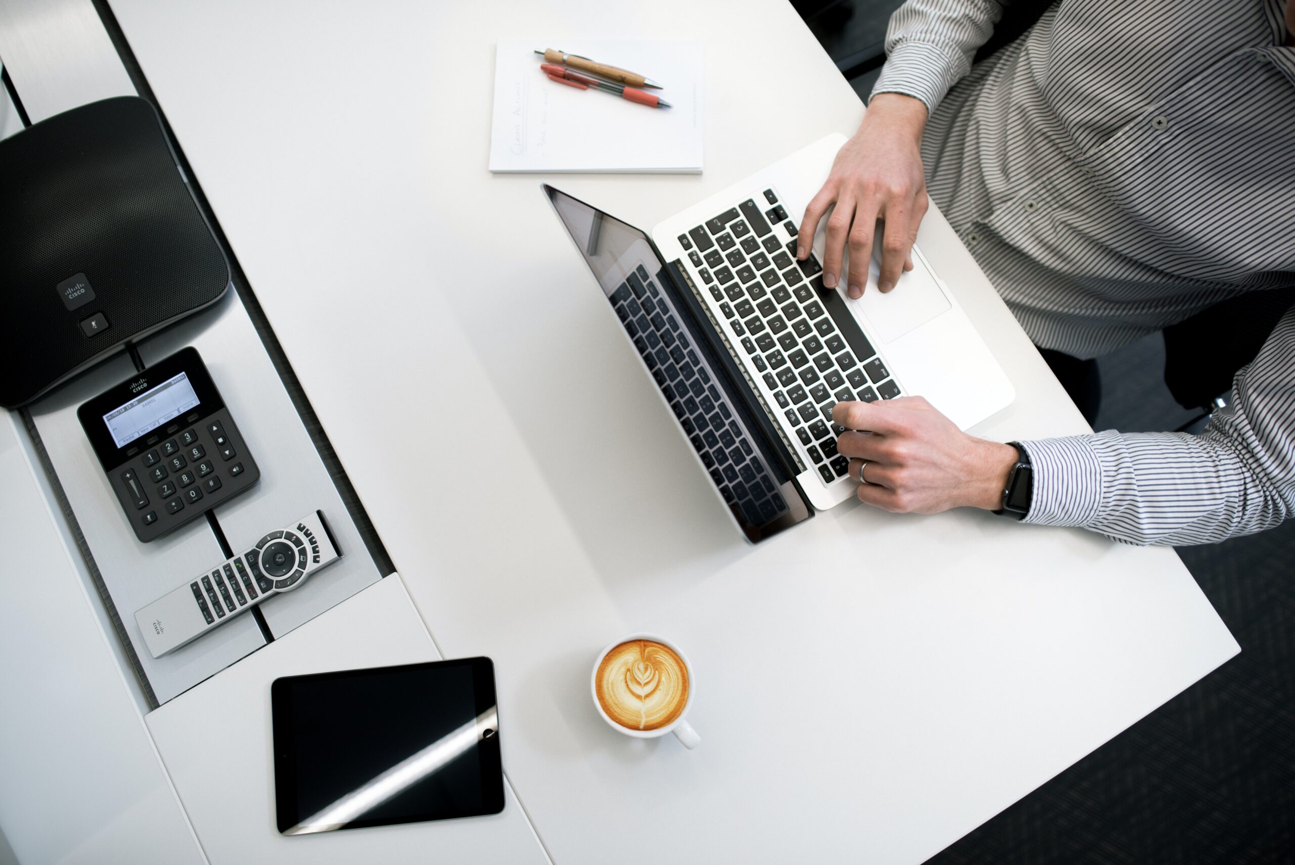 Person working on a laptop at a white desk with a coffee cup, notepad, pens, a tablet, a phone, and a printer nearby.
