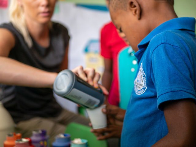 A child in a blue shirt pours liquid from a container into a cup, assisted by an adult, with art supplies visible on the table.