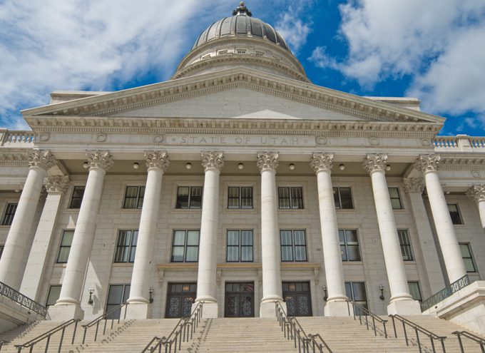 Front view of the Utah State Capitol building with tall columns, large dome, and wide steps, set against a partly cloudy blue sky.