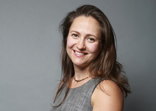 A woman with long brown hair, wearing a gray sleeveless top and a necklace, smiles at the camera against a plain gray background.