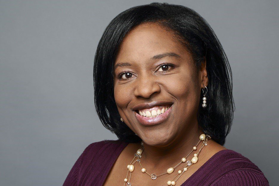 A woman with straight black hair smiles at the camera, wearing a purple top and a pearl necklace against a plain gray background.