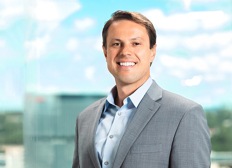 A man in a gray suit and light blue shirt smiles while standing in front of a blurred cityscape background.