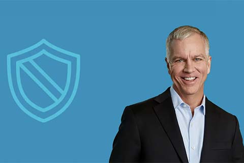 Smiling man in a dark suit and light blue shirt stands in front of a blue background with a shield icon on the left.