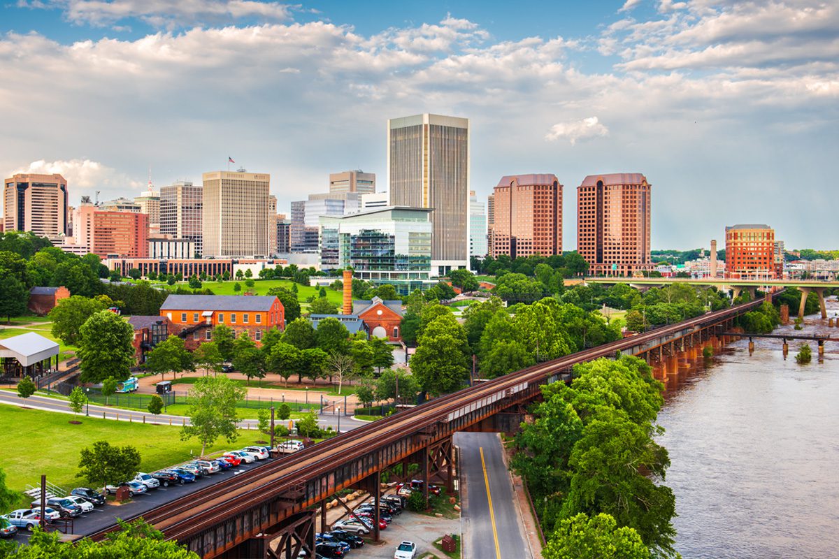 A city skyline with modern and historic buildings, a river in the foreground, and a railway bridge crossing over, surrounded by trees and a parking lot.