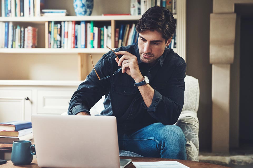 A man sits in a living room holding glasses and a pen, looking at a laptop on a table with bookshelves in the background.