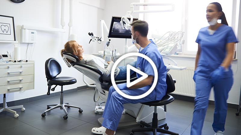 A dentist examines a patient in a dental clinic while another staff member walks past; dental equipment and monitors are visible in the room.