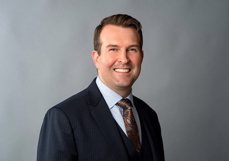 A man in a dark suit, light blue shirt, and patterned tie smiles at the camera against a plain gray background.