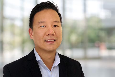 A man wearing a dark blazer and light shirt stands indoors in front of a blurred background with large windows.