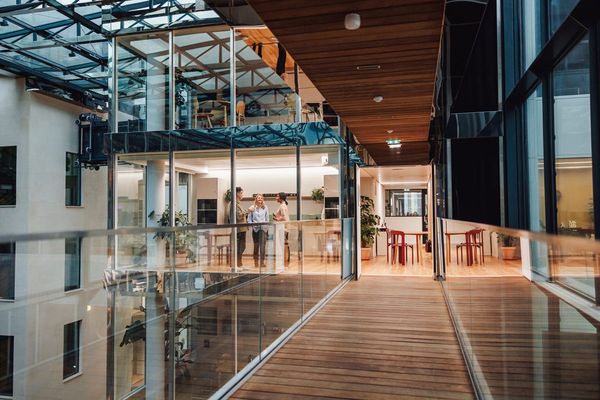 A group of professionals engage in discussion while standing in a well-lit office café, showcasing modern business attire and the setting’s sleek design.