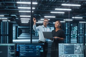 Data Center Black Female e-Business Entrepreneur and White Male IT Specialist talk, Use Laptop. Information Technology Engineer and System Administrator work in Big Cloud Computing Server Farm.