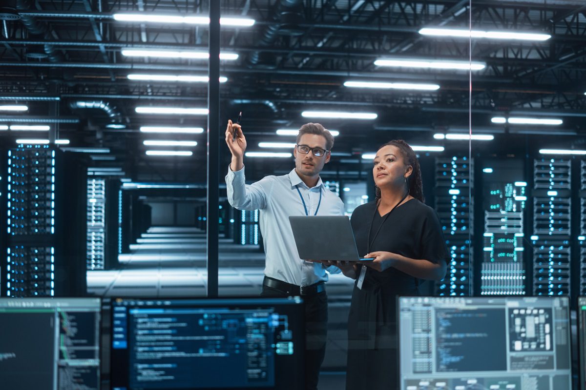 Data Center Black Female e-Business Entrepreneur and White Male IT Specialist talk, Use Laptop. Information Technology Engineer and System Administrator work in Big Cloud Computing Server Farm.