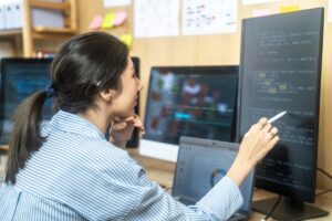 Focused young software developer debugging code on screens in modern workspace surrounded by technology. Depicts problem solving and software development skills.