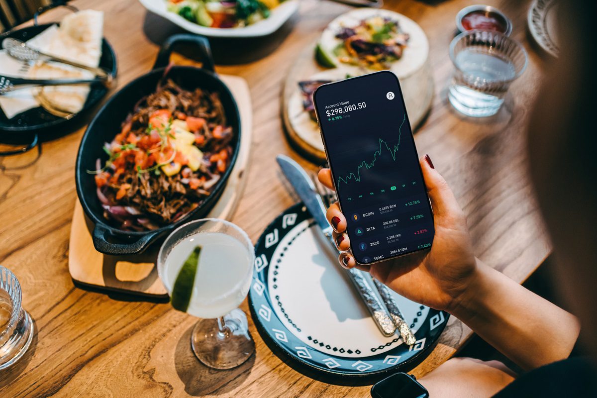 Young Asian woman managing personal finance and investment. Analyzing investment and stock market trades via mobile app in restaurant. Making financial plans and setting financial goals with smartphone. Wealth management and online banking concept