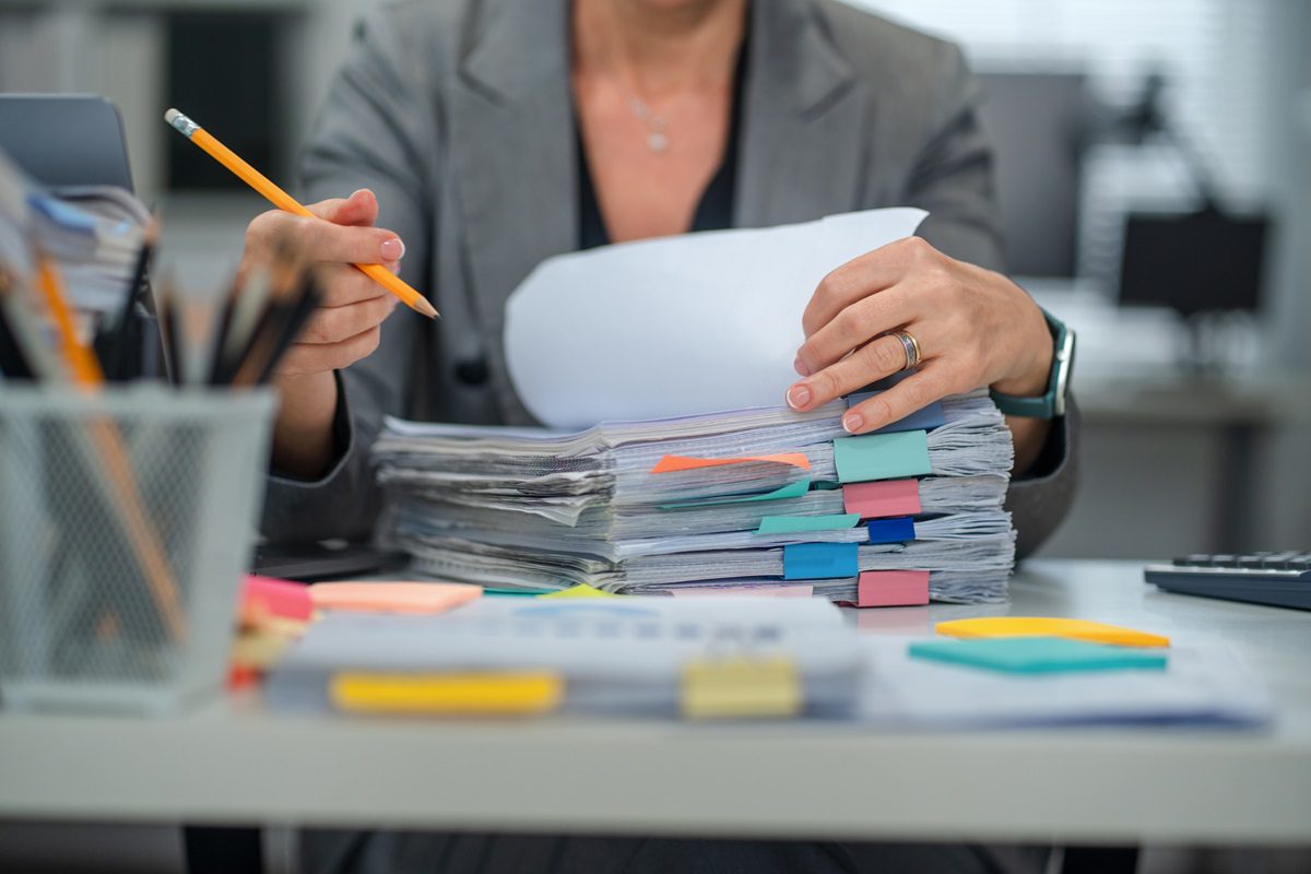Senior businesswoman, searching documents in messy stack. Reports, forms, file folders, ordering and organization. Frustration, emotional stress, business office overflow.