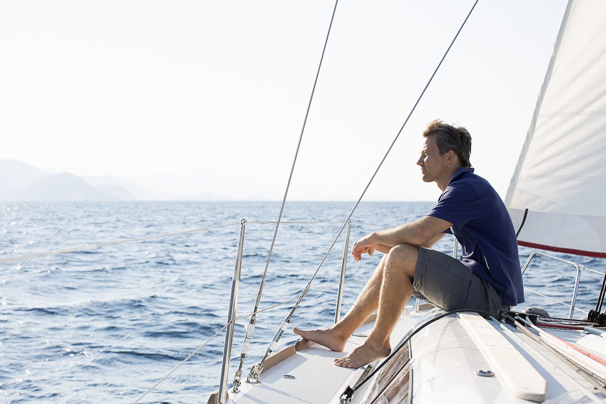 A man sits barefoot on the deck of a sailboat, looking out at the open sea on a clear day.