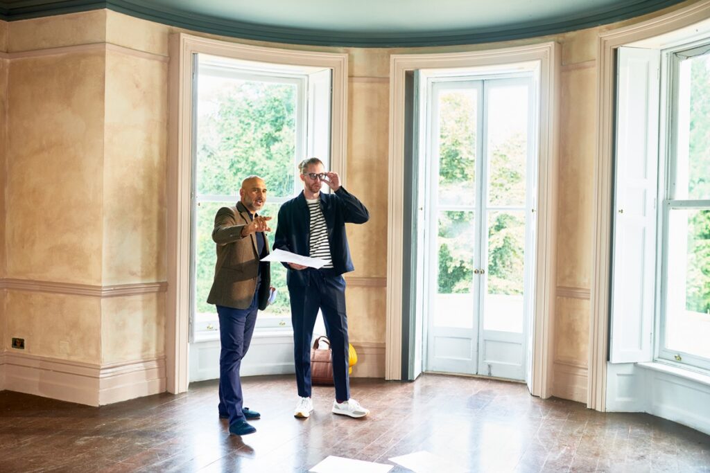 Building developer standing in spacious empty room of house talking to mature man about potential development plans, planning, expertise, development