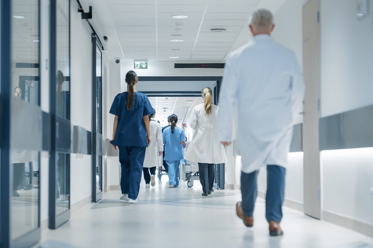 Hospital Hallway with Doctors, Nurses and Specialists in Hospital. Female and Male Physicians, Surgeons, Healthcare Officials Walk Together in Corridor with Their Back to Camera