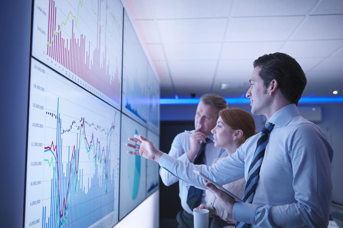 Three business people discuss graphs on screen in meeting room