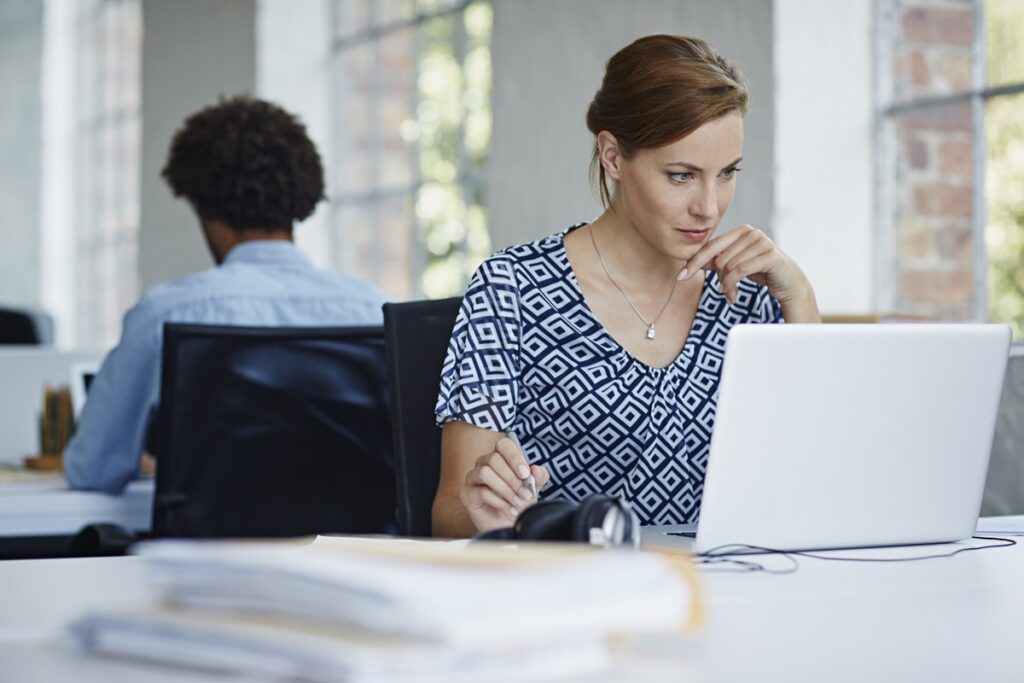 Woman focused on a laptop at her office desk with a stack of documents, reviewing compliance paperwork