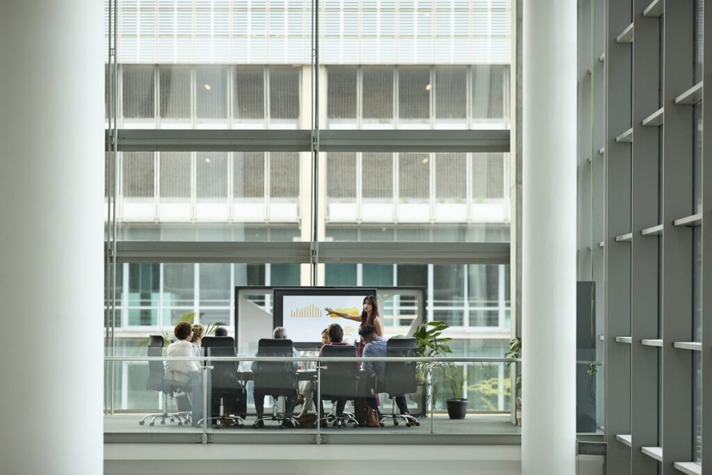 A woman presents a bar chart on a digital screen to colleagues seated at a conference table in a glass-walled meeting room.