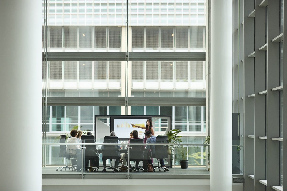 A woman presents a bar chart on a digital screen to colleagues seated at a conference table in a glass-walled meeting room.