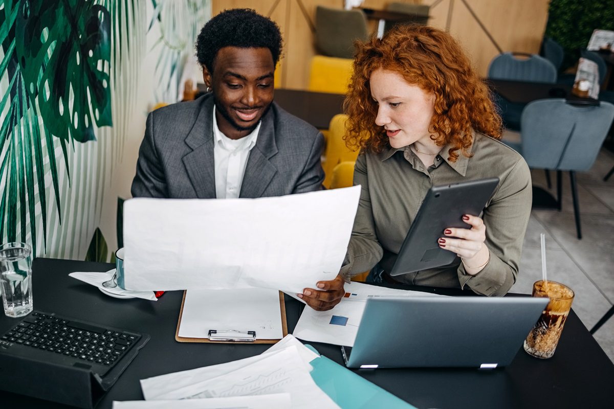 Two colleagues review printed documents and a tablet together at a desk covered with paperwork and a laptop in a modern office