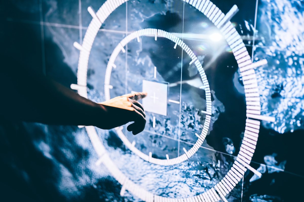 A hand of an unrecognizable person reaching toward and interacting with a glowing digital interface, symbolizing the future of human-computer interaction, advanced analytics, and technological innovation. The circular design interface with intricate data visualizations illustrates the concepts of space exploration, cybersecurity, military defence, satellite locating and geofencing, and augmented reality applications. Modernity, science fiction, and digital transformation