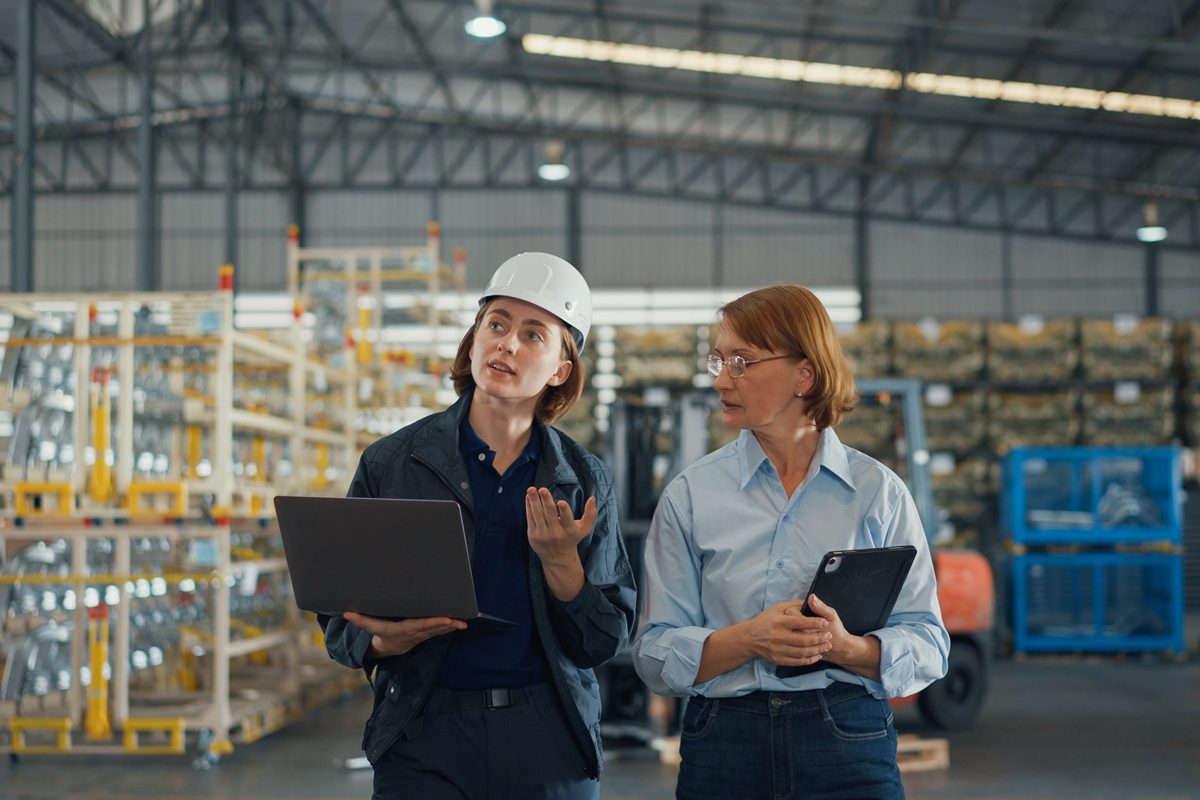 Young caucasian woman worker and mature female manager walking checks stock inventory with tablet and laptop and discuss talk together in retail warehouse. Logistic industry business concept.