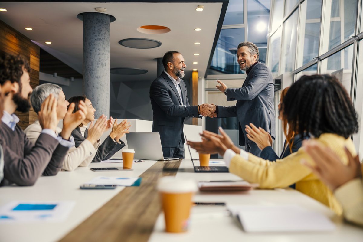 Business leaders shaking hands, celebrating a successful partnership and deal, while smiling diverse colleagues are clapping at the modern office meeting