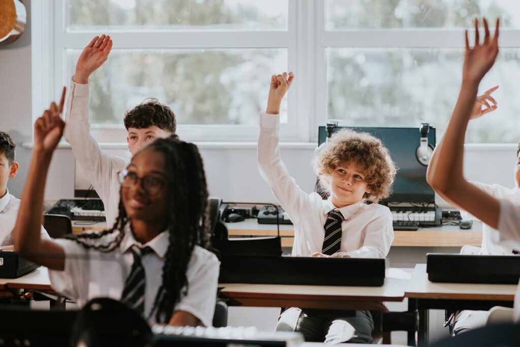 A boy raises his hand in a classroom setting. He wears a school uniform and computer equipment is visible in the background. Depicts education.